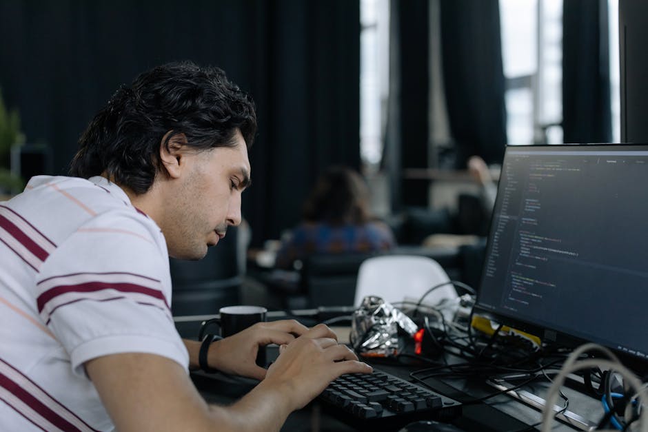 A young man with dark, wavy hair is seated at a desk, focused on typing on a keyboard while working on a desktop computer with a large monitor displaying lines of code. The workspace is indoors with l